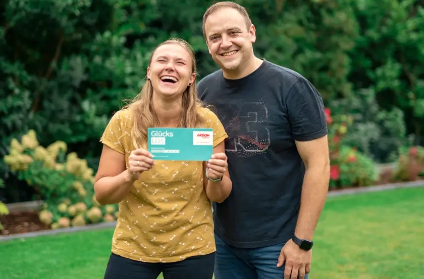 Die Gewinner*innen Bozena und Sebastian stehen in sommerlicher Kleidung nebeneinander in ihrem Garten. Bozana hält ihr Glückslos in die Kamera und lacht.