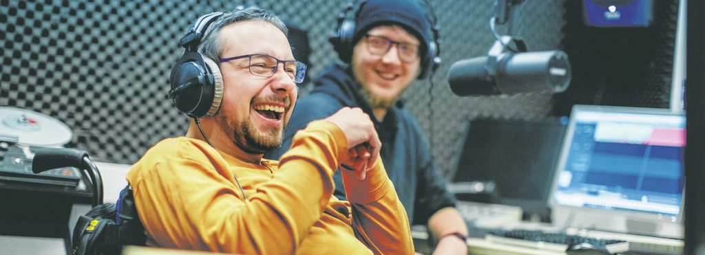 Ein Mann im Rollstuhl sitzt lachend in einem Radiostudio vor einem Mikro. Er hat große Kopfhörer auf, trägt einen Vollbart, Brille und einen dunkelgelben Pulli. Im Hintergrund sitzt ein zweiter Mann vor einem Computerbildschirm. Er guckt lachend zu dem Mann im Rollstuhl. Er ist jünger, hat ebenfalls einen Vollbart und eine Brille. Über seiner schwarzen Mütze hat auch er große Kopfhörer aufgesetzt.