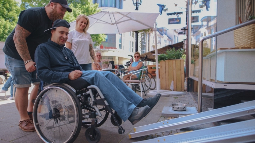 Ein junger Mann hilft einem jungen Mann im Rollstuhl eine Rampe hinauf. Daneben steht eine junge Frau und schaut zu.