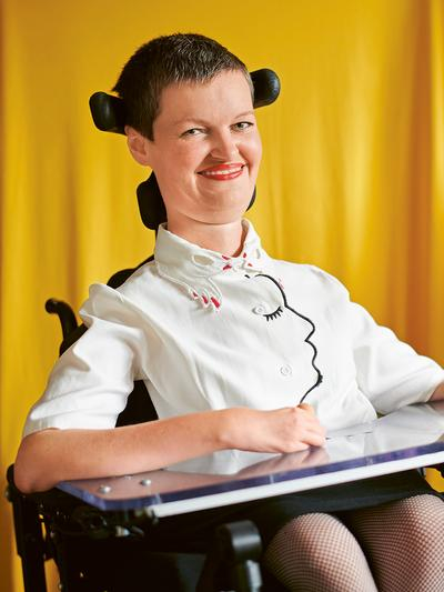 eine junge Frau mit kurzem Haar und weißer Bluse im E-Rollstuhl