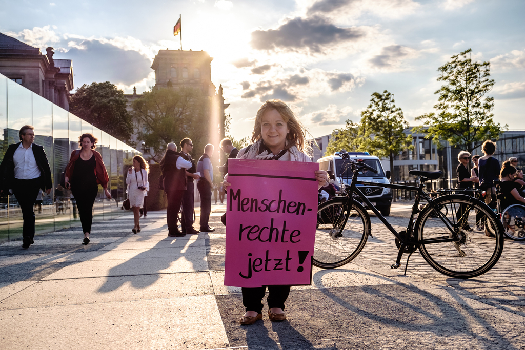 Eine kleinwüchsige Frau hält im Abendlicht ein Plakat mit der Forderung: Menschenrechte jetzt!