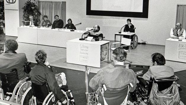 Mehrere Personen im Rollstuhl verfolgen eine Diskussionsveranstaltung 