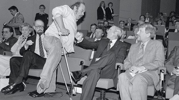 Ein Mann mit Krücken bedroht einen sitzenden Mann, der bei einer Veranstaltung in der ersten Reihe sitzt.