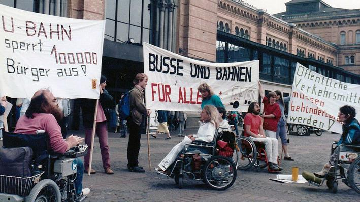 Mehrere Rollstuhlfahrer und andere Personen protestieren mit Plakaten vor einem Gebäude
