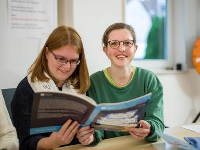 Zwei Frauen sitzen an einem Schreibtisch und halten ein Buch in den Händen, die linke Frau liest, die rechte Frau lächelt in die Kamera.