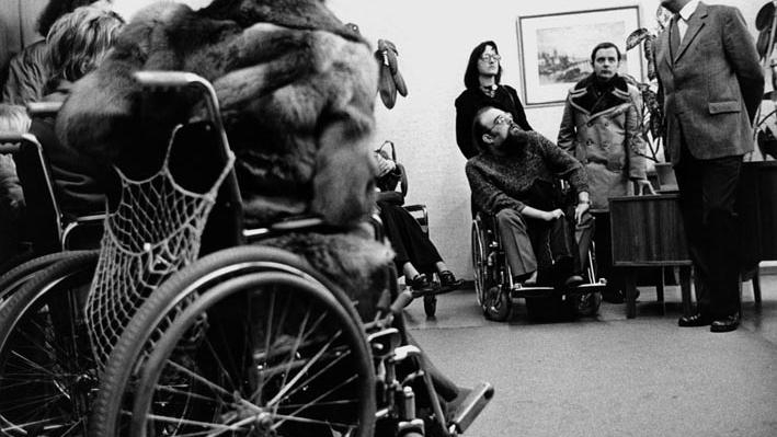 Eine Gruppe von Menschen, viele davon im Rollstuhl, im Gespräch mit einem Mann im Anzug