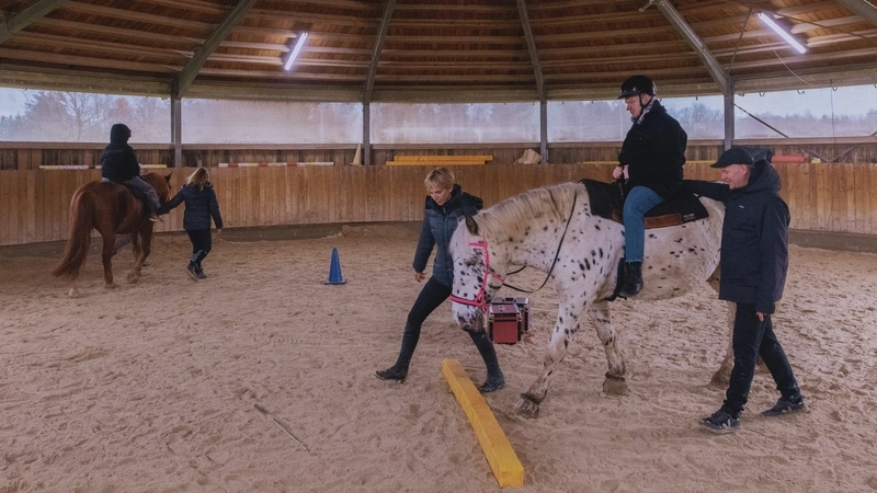 In einer Reithalle führt eine Frau einen Schimmel am Halfter im Kreis. AUf dem Pferd sitzt ein älterer Mann mit Reithelm. Er wird gesichert von einem zweiten Begleiter, der neben dem Pferd herläuft.