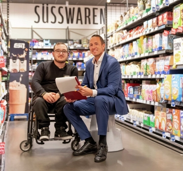 In einem Supermarkt-Gang zwischen zwei Regalreihen sitzen zwei Männer. Links ein Mann in schwarzer Hose und Pulli im Rollstuhl mit Brille und mit Bart, rechts neben ihm ein Mann mit kurzen dunklen Haaren in blauem Anzug und weißem Hemd auf einem Tritthocker. Beide lächeln in die Kamera.