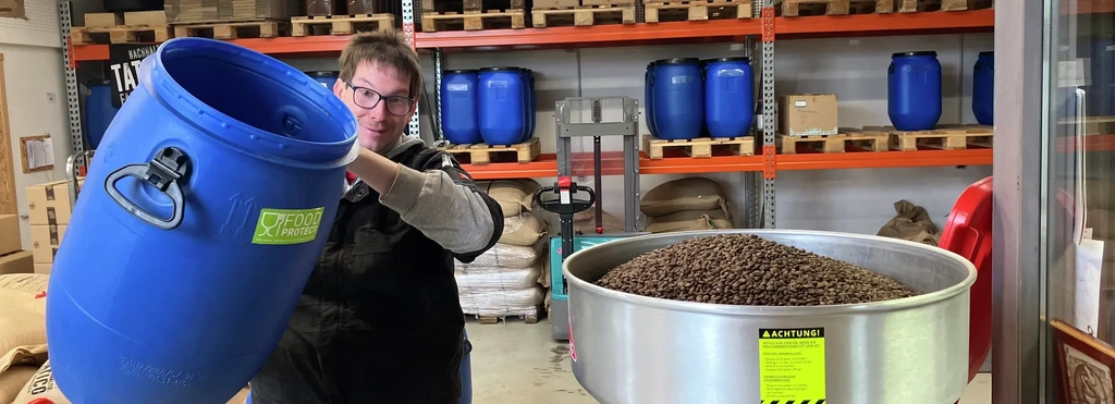 Ein Menn mit Brille schüttet Kaffeebohnen aus einer großen blauen Tonne in einen Trichter