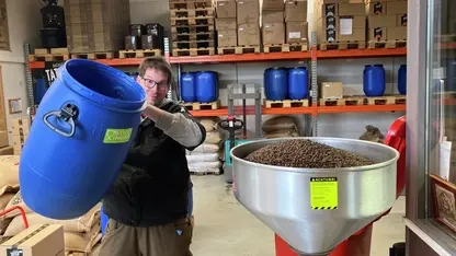 Ein Menn mit Brille schüttet Kaffeebohnen aus einer großen blauen Tonne in einen Trichter