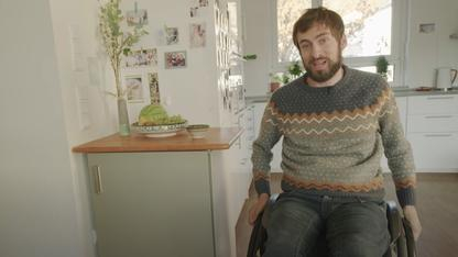 In einer modern eingerichteten Küche ist ein junger Mann im Rollstuhl zu sehen, der einen gemusterten Pullover trägt und in die Kamera schaut.
