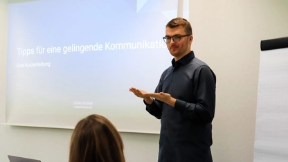 Heiko Burak, ein junger Mann mit Brille und kurzen braunen Haaren, steht vor einer Leinwand auf der "Tipps für eine gelingende Kommunikation" steht. Er gebärdet.