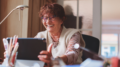 Eine Frau mit kurzen roten Haaren und Brille sitzt an einem Schreibtisch und hat ein Tablet in der Hand und lacht in die Richtung des Tablets.