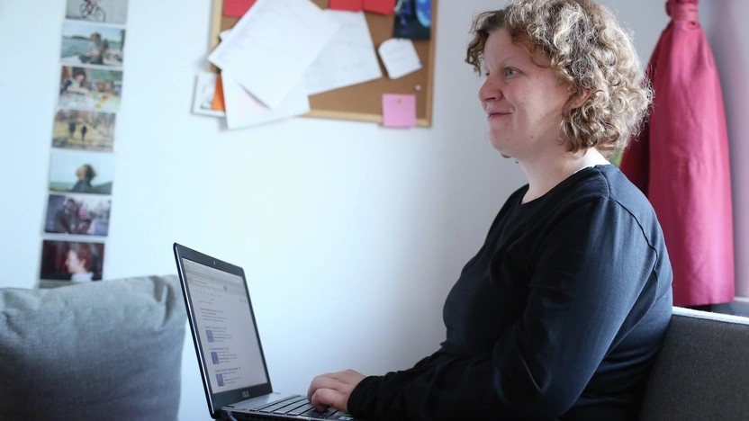 Eine Frau mit kurzen lockigen Haaren sitzt auf einem Sofa. Sie hat einen Laptop auf dem Schoß stehen.