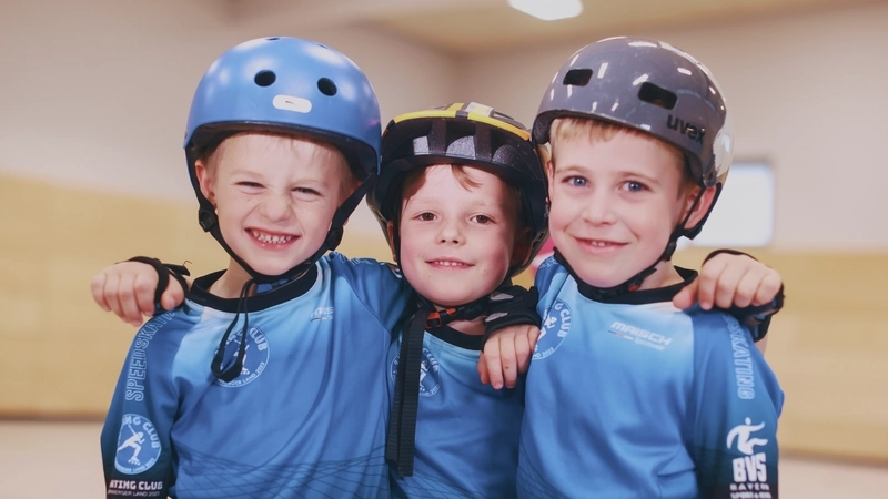 Drei kleine Jungs im Grundschulalter stehen Arm in Arm dicht beieinander in einer Sporthalle. Sie alle tragen hellblaue Sporttrikots mit dem Vereinslogo des "Speed-Skating-Club Nürnberger Land" und Sturzhelme.