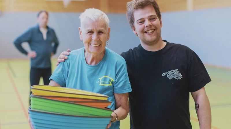 Eine Seniorin mit kurzen weißen Haaren und hellblauem T-Shirt steht in einer Turnhalle neben einem jungen Mann mit kurzen dunkelblonden Haaren in dunklem T-Shirt, der ihr seinen rechten Arm um die Schulter gelegt hat. Die alte Frau hält bunte Plastikhütchen in ihren Händen.