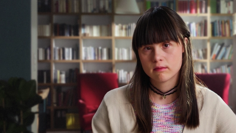 Eine junge Frau mit Down-Syndrom sitzt in einer Wohnung. Im Hintergrund sieht man ein Wohnzimmer mit Bücherregal. Sie schaut ernst in die Kamera.