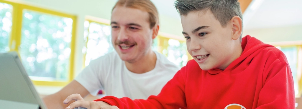 Ein Junge und ein junger Erwachsener sitzen nebeneinander und schauen auf einen Laptop. Vor ihnen liegt ein Controller.