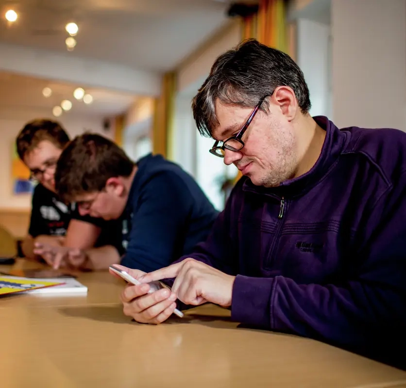 Ein Mann sitz an einem Tisch und schaut auf ein Smartphone. Neben ihm am Tisch sitzen zwei junge Männer, die gemeinsam auf ein weiteres Smartphone gucken.