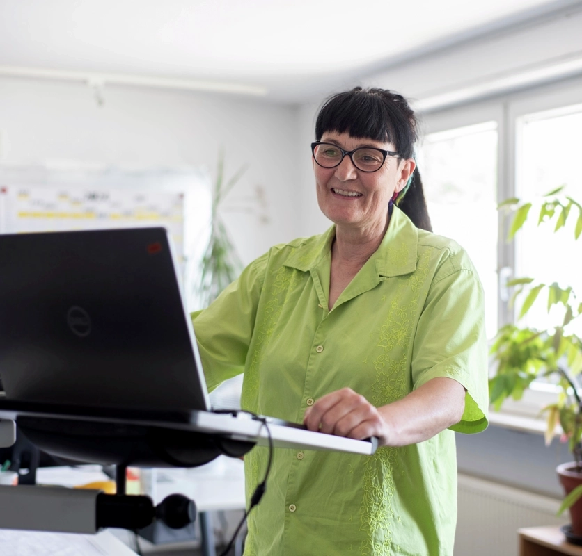 Eine Frau steht in einem Büroraum an einem Laptop. Sie trägt einen Brille, einen Pferdeschwanz und eine hellgrüne, kurzärmelige Bluse.