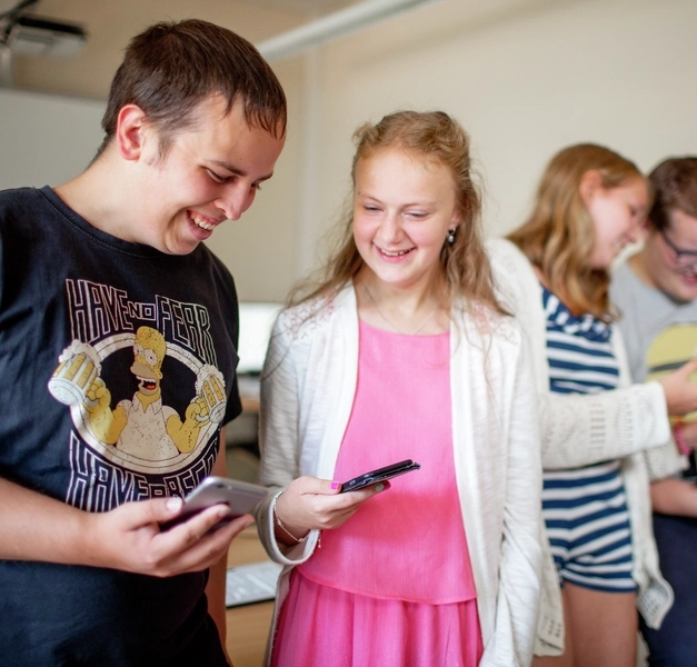 Zwei Jugendliche stehen nebeneinander und schauen lachend auf ihre Smartphones.