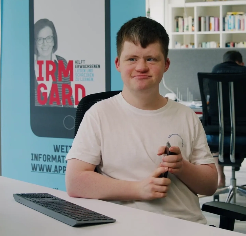 Eine Person mit Behinderung sitzt in einem offenen Büro am Schreibtisch und schaut auf einen Bildschirm.