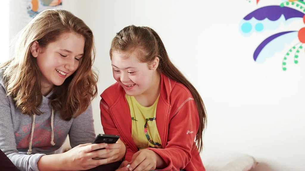Zwei Jugendliche sitzen neben einander auf einem Bett und schauen auf ein Handy. Eine von ihnen hat das Down-Syndrom.