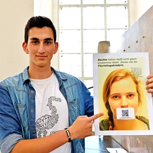 Talha hält eines seiner Plakate in der Hand, auf der ein Mädchen mit QR-Code anstelle eines Mundes zu sehen ist.