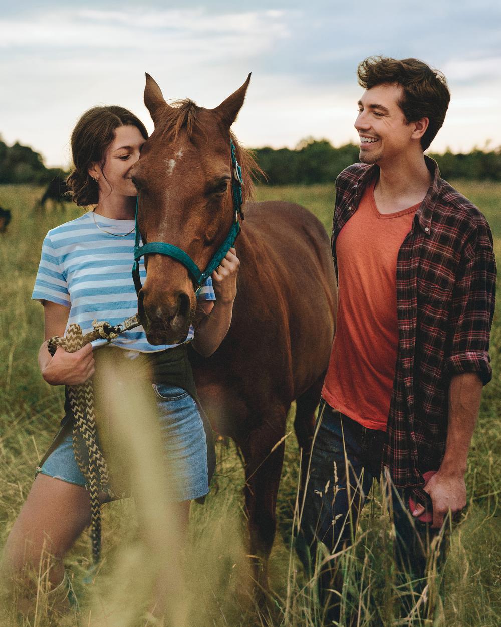 Auf einer großen Pferdeweide stehen eine junge Frau und ein junger Mann. Zwischen ihnen steht ein braunes Pferd, das die Frau am Halfter festhält.