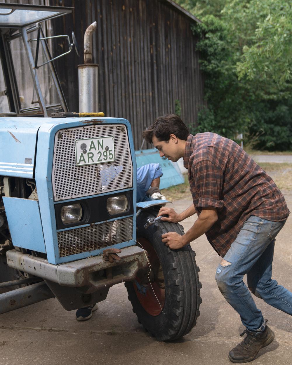 Ein junger Mann mit kariertem Flanellhemd steht vor einem alten, blauen Traktor. Er hat beide Hände auf einen der Vorderreifen gelegt und stemmt sich kraftvoll dagegen.