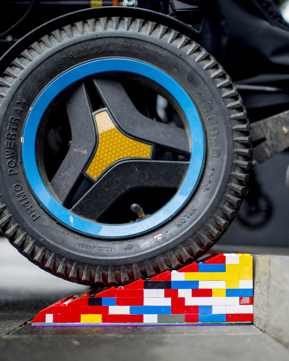 Eine Legorampe vor einer Stufe. Im Anschnitt ist der Reifen eines Elektrorollstuhls, der über die Rampe rollt.