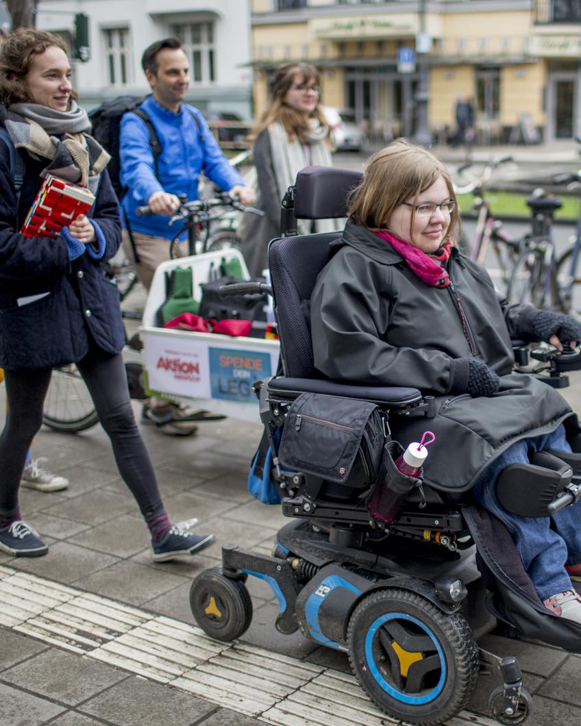 Ein Gruppe junger Leute ist gemeinsam in Köln unterwegs. Eine junge Frau benutzt einen Elektrorollstuhl. Ein Mann im Hintergrund schiebt ein Lastenrad. Eine andere junge Frau hält eine Rampe aus Legosteinen in der Hand.