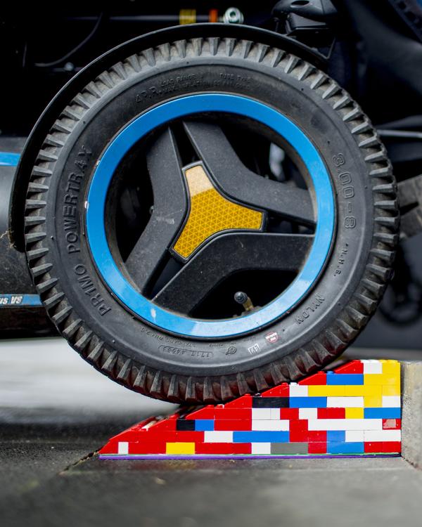 Das runde Bild eines Rollstuhlreifens, der über eine Rampe aus Legosteinen fährt. Um das Bild herum sind geschwungene, rote Linien zu sehen, die das Abspielen einer Audiodatei andeuten.