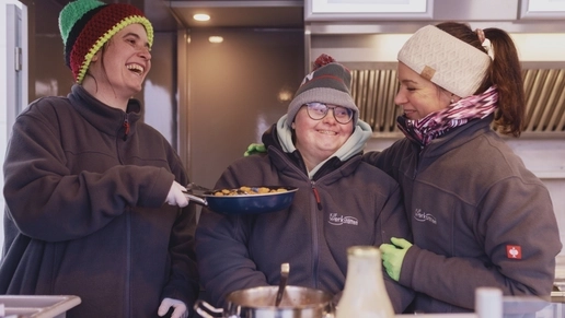 Drei Frauen stehen in einem Foodtruck und lachen. Alle drei tragen Mützen und einheitliche graue Jacken. Die Frau in der Mitte hat eine sichtbare Behinderung. Die Frau links neben ihr hält eine Pfanne mit Essen drin hoch. Die Frau auf der anderen Seite legt der mittleren Frau den Arm um die Schulter.