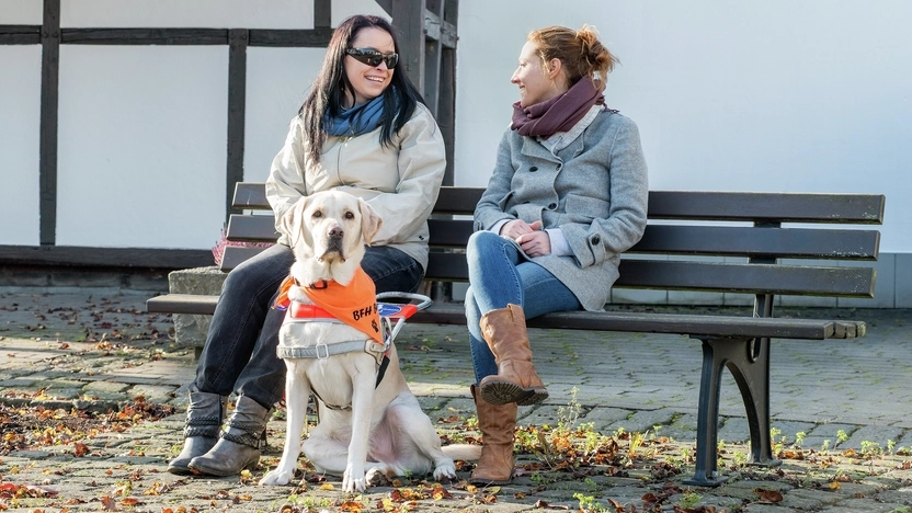 Zwei junge Frauen in warmen Jacken sitzen auf einer Bank, die vor einem Fachwerkhaus steht. Eine der beiden hat einen Blindenhund dabei und eine dunkle Brille auf. Sie reden miteinander.