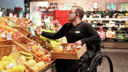 In einem Supermarkt-Gang prüft ein Mitarbeiter in schwarzer Hose und Pulli, der im Rolli sitzt,  Obst an einem Obststand. Aussortierte Ware legt er in einem Korb auf seinem Schoß.