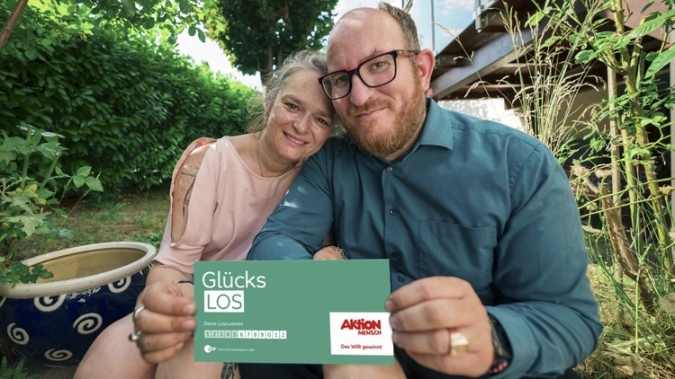 Ein Paar sitzt zusammen im Freien, sie lächeln freundlich. Sie halten ein Glücks-Los der Aktion Mensch in der Hand. Die Atmosphäre wirkt entspannt und freundlich.