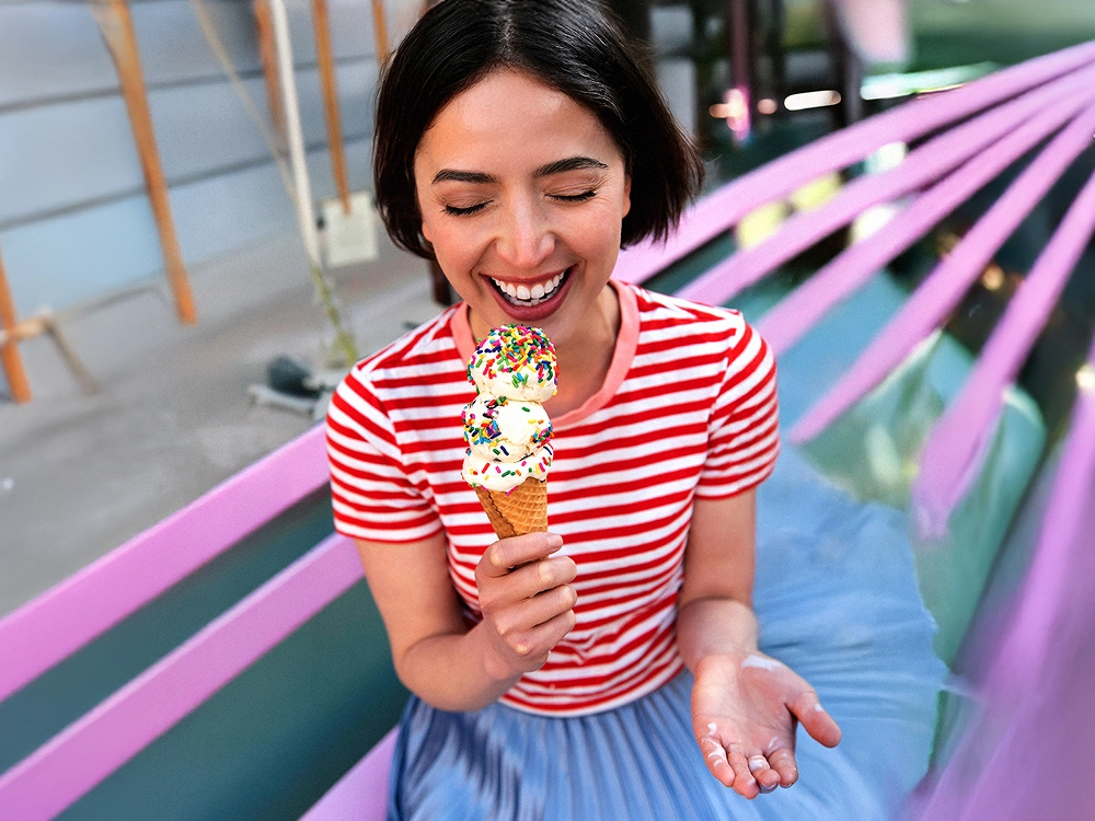 Eine junge Frau mit mittellangen, dunklen Haaren im rot-weiß-gestreiften Shirt lacht glücklich. In der Hand hält sie eine Eiswaffel mit drei Kugeln Vanille-Eis, die mit bunten Zuckerstreuseln bestreut sind.