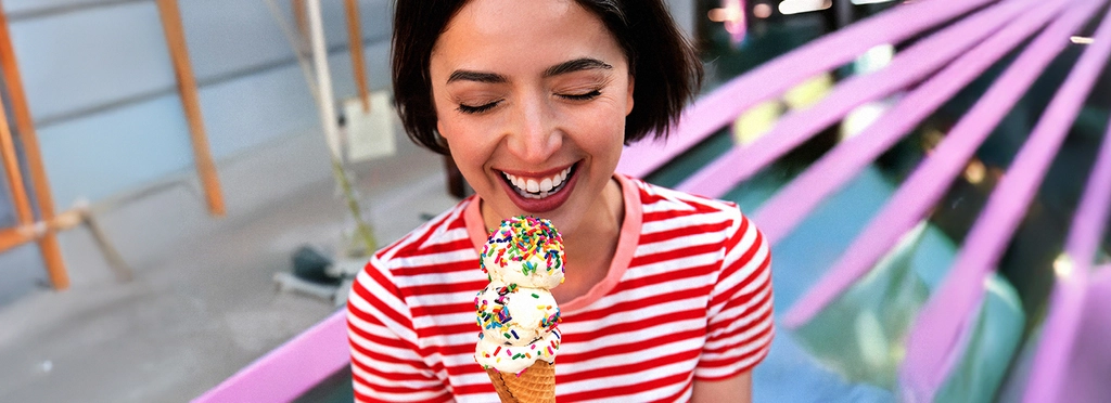 Eine junge Frau mit mittellangen, dunklen Haaren im rot-weiß-gestreiften Shirt lacht glücklich. In der Hand hält sie eine Eiswaffel mit drei Kugeln Vanille-Eis, die mit bunten Zuckerstreuseln bestreut sind.