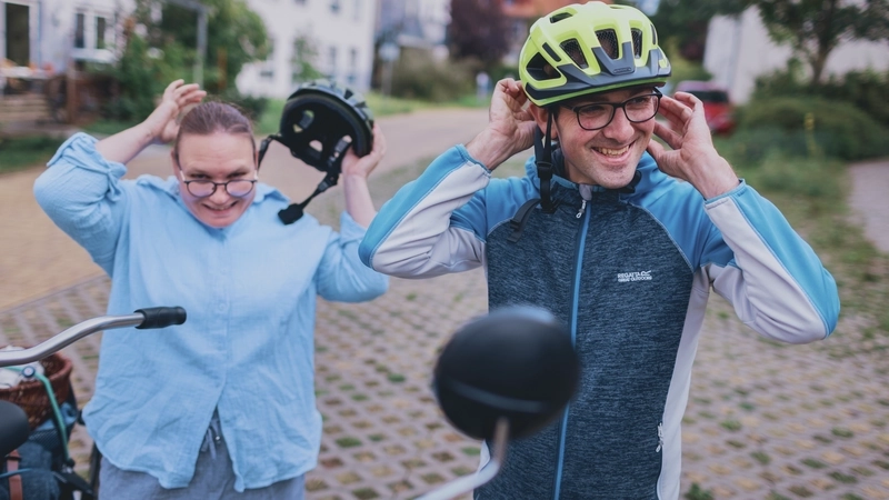 Eine Frau und ein Mann in hellblauer Freizeitkleidung stehen auf einer Freifläche vor Wohnhäusern und setzen ihre Fahrradhelme auf. Vor ihnen sieht man die Lenkergriffe eines Tandems.