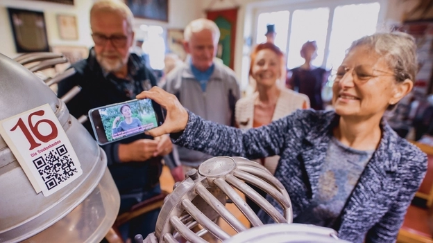 Eine Gruppe Besucher*innen steht an einem alten Frisierstuhl. Über ein Mobiltelefon schauen sie sich ein Gebärdensprachvideo zum Exponat an.