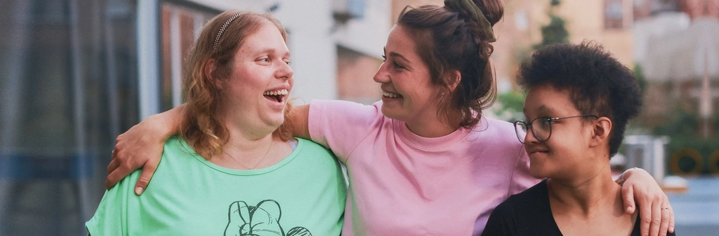 Drei Frauen stehen nebeneinander und haben jeweils einen Arm umeinander gelegt. Sie lachen.