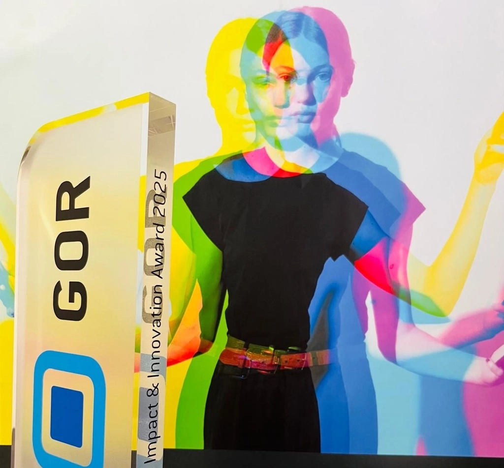 Das Bild zeigt den gläsernen Pokal des GOR Impact and Innovation Awards 2025. Im Hintergrund ist ein Ausschnitt des Veranstaltungsplakats zu sehen.