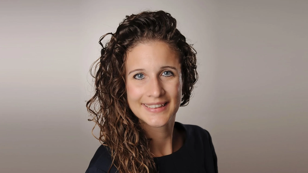 Porträt von Lea Thönnes - einer Frau mit langem lockigen Haar vor einem neutralen Hintergrund. Sie lächelt.