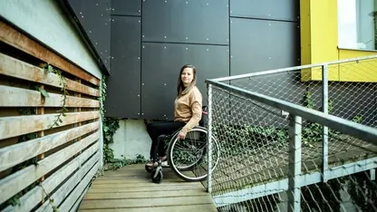 Eine junge Frau im Rollstuhl fährt eine Rampe an einem Haus runter.