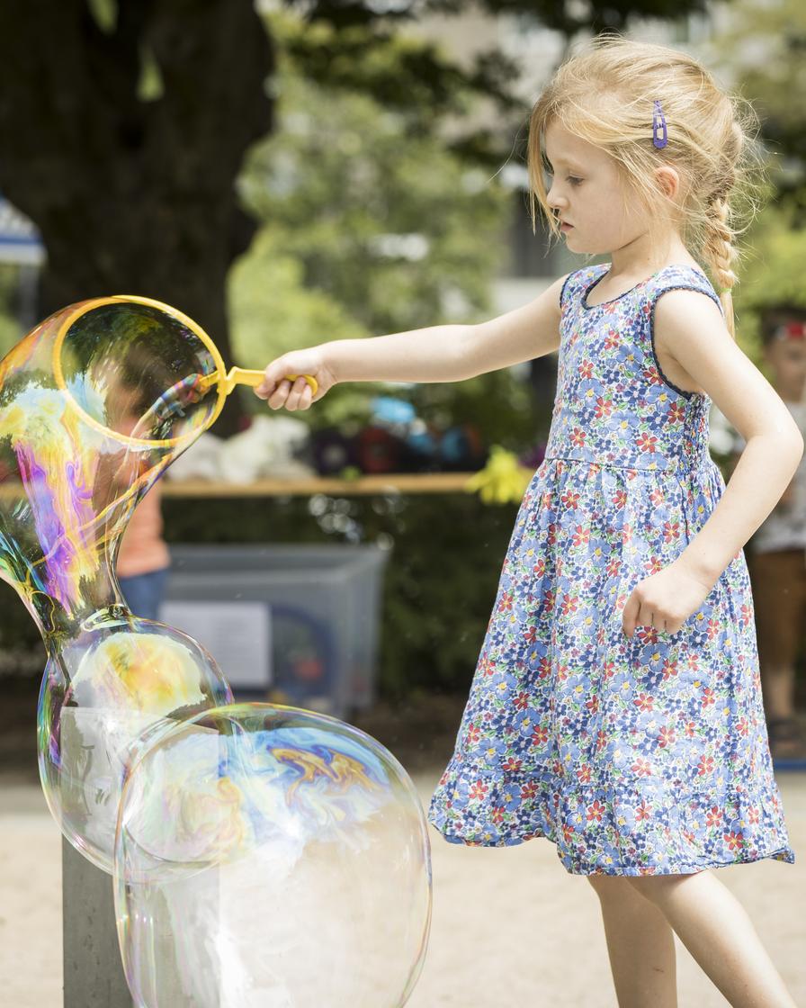 Auf einem Kinderspielplatz steht ein blondes Mädchen in einem geblümten Kleid. In der rechten Hand hält sie ein Spielzeug, mit dem sie große, bunte Seifenblasen macht.