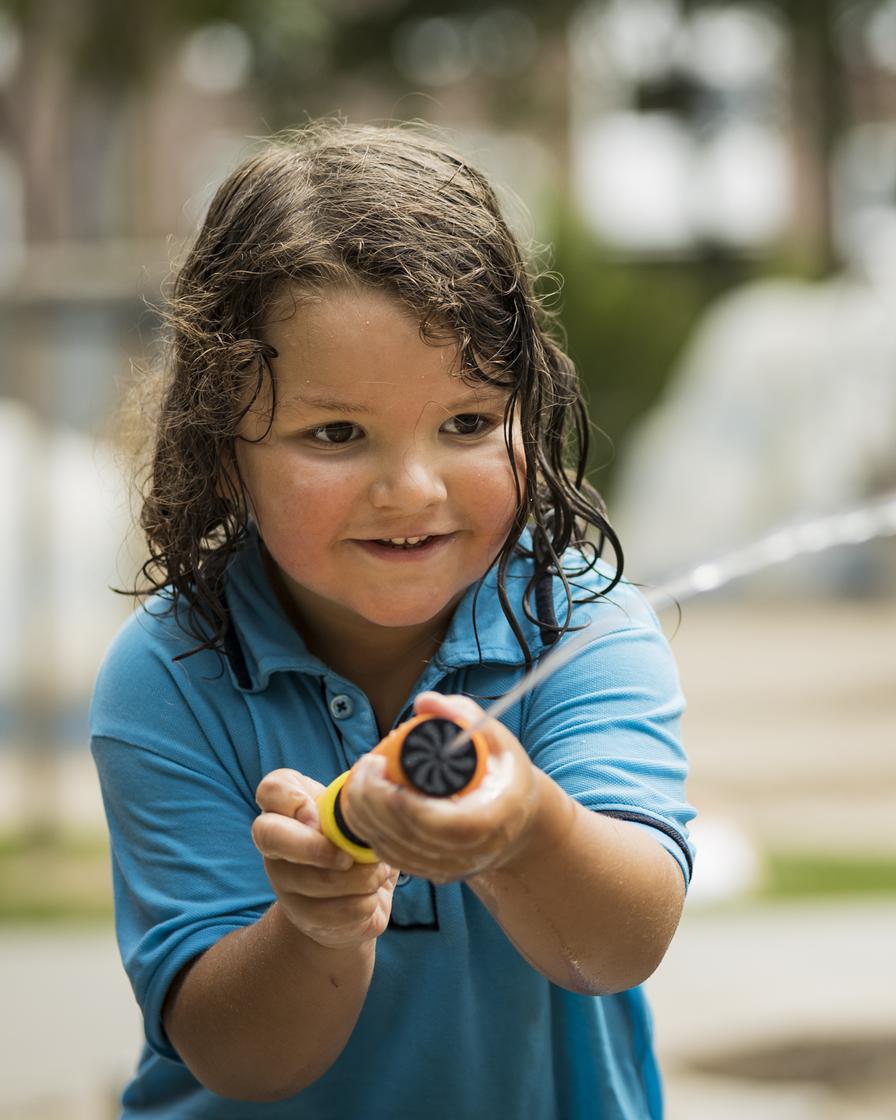 Auf einem Spielplatz ist ein Kind mit längeren braunen Haaren zu sehen. Die Haare sind leicht nass. In den Händen hält das Kind einen Wasserschlauch und spritzt damit Wasser nach vorne weg.