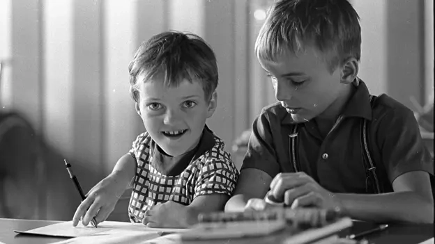 Ein Mädchen mit verkürzten Armen schreibt mit einem Stift auf einem Papier. Ein Junge sitzt neben ihr.