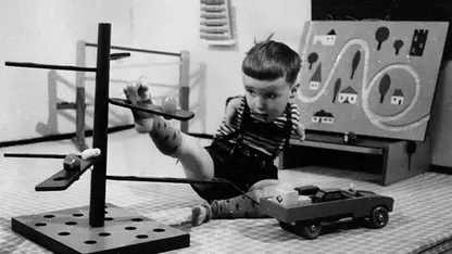 ein kleines Kind mit verkürzten Armen hebt mit seinem Fuß Bauklötze auf ein Gestell.