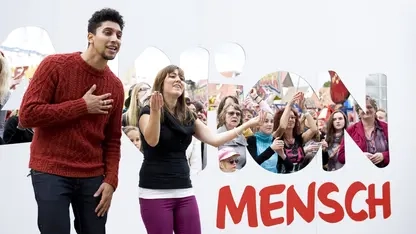 Andreas Bourani und Lisa tanzen vor dem Logo der Aktion Mensch. Im Hintergrund sind lachende Menschen zu sehen.